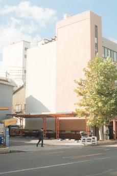 A man in a black suit is walking on the sidewalk next to a parking lot with a large building in the background. The parking lot is mostly empty and partially covered by a roof structure held up by orange beams. There is a large tree with green leaves to the right of the parking lot. The building in the background is modern, with a light pastel façade, and there are additional buildings in the background. The sky is clear with a few clouds.
