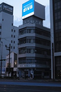 A multi-story building with a large, illuminated sign on top displaying 'Pachinko & Slot Divo'. The building appears to have a modern architectural style with glass windows and a gray facade. It's situated in an urban environment, with other buildings visible in the background. The scene is set during a time of low lighting, suggesting early morning or evening, as indicated by the dark sky and street lights.