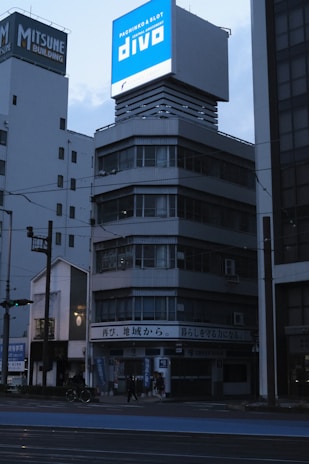 A multi-story building with a large, illuminated sign on top displaying 'Pachinko & Slot Divo'. The building appears to have a modern architectural style with glass windows and a gray facade. It's situated in an urban environment, with other buildings visible in the background. The scene is set during a time of low lighting, suggesting early morning or evening, as indicated by the dark sky and street lights.