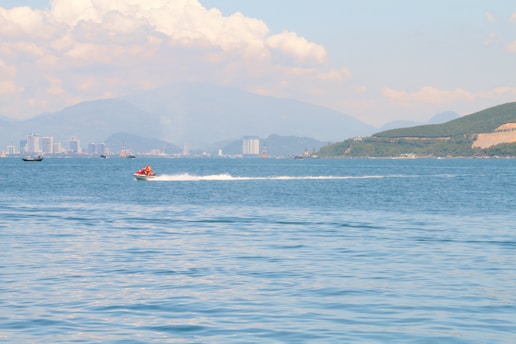A person rides a jet ski across a calm body of water, creating a white wake behind. In the background, there are distant city buildings and mountainous terrain under a partly cloudy sky.