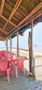 A rustic beachside setup with a wooden structure featuring a thatched roof, overlooking the sea. Inside the structure, several red plastic chairs surround a table. The view extends out to the sandy beach and the ocean, creating a tranquil and scenic atmosphere.