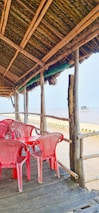 A rustic beachside setup with a wooden structure featuring a thatched roof, overlooking the sea. Inside the structure, several red plastic chairs surround a table. The view extends out to the sandy beach and the ocean, creating a tranquil and scenic atmosphere.