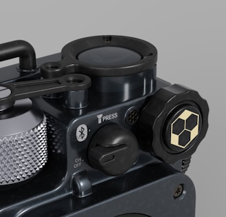 A close-up view of a mechanical device with various components such as a textured silver dial, a Bluetooth symbol, and a button marked 'PRESS'. The surface is dark with a metallic finish, featuring a hexagonal design on a knob. The overall appearance is sleek and industrial.
