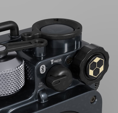 A close-up view of a mechanical device with various components such as a textured silver dial, a Bluetooth symbol, and a button marked 'PRESS'. The surface is dark with a metallic finish, featuring a hexagonal design on a knob. The overall appearance is sleek and industrial.