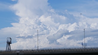 Large, billowing white clouds dominate the sky above a concrete bridge. The bridge features railings and two tall lampposts, with a small structure possibly for maintenance or observation on the left side. The sky is a bright blue.