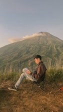 A person sits on a foldable chair, reading a book against the backdrop of a mountain with clouds drifting over the peak. The ground is earthy with sprouting grass, and there's a small visible corner of a tent.