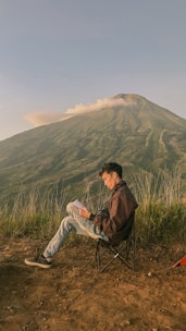 A person sits on a foldable chair, reading a book against the backdrop of a mountain with clouds drifting over the peak. The ground is earthy with sprouting grass, and there's a small visible corner of a tent.
