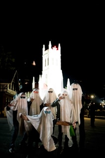 A group of people dressed as ghosts stand in front of a brightly illuminated church at night. Each person wears a white sheet with sunglasses, adding a playful touch to the spooky theme. The dark night contrasts with the well-lit church, creating a visually striking scene.