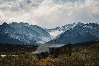 A rustic cabin nestled in a landscape of tall snow-capped mountains and surrounded by lush green foliage. The cloudy sky adds a dramatic backdrop, enhancing the sense of tranquility.