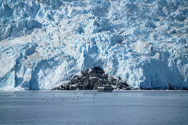 A vast glacier dominates the scene, its massive ice formations cascading down towards the ocean water, reflecting a pale blue hue. In front of the glacier, a small boat traverses the icy waters, providing a sense of scale to the immense natural structure. A flock of birds can be seen flying low over the water, adding a touch of life to the cold, imposing environment.