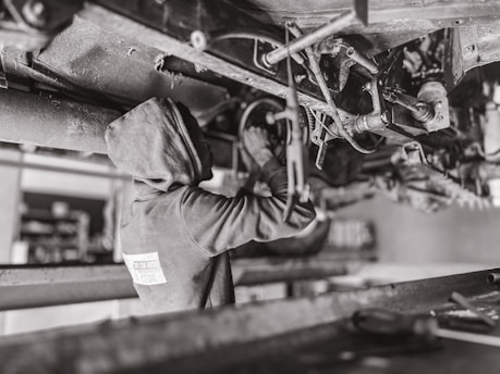 A mechanic wearing a hooded sweatshirt works under a vehicle, performing maintenance or repairs. The setting is likely a workshop or garage. The scene is in black and white, emphasizing the components and mechanical parts of the vehicle's underside.