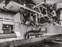 A mechanic wearing a hooded sweatshirt works under a vehicle, performing maintenance or repairs. The setting is likely a workshop or garage. The scene is in black and white, emphasizing the components and mechanical parts of the vehicle's underside.