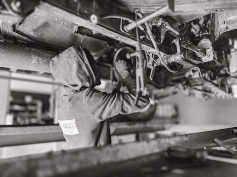 A mechanic wearing a hooded sweatshirt works under a vehicle, performing maintenance or repairs. The setting is likely a workshop or garage. The scene is in black and white, emphasizing the components and mechanical parts of the vehicle's underside.