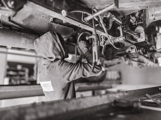 A mechanic wearing a hooded sweatshirt works under a vehicle, performing maintenance or repairs. The setting is likely a workshop or garage. The scene is in black and white, emphasizing the components and mechanical parts of the vehicle's underside.