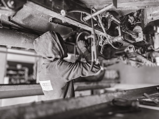A mechanic wearing a hooded sweatshirt works under a vehicle, performing maintenance or repairs. The setting is likely a workshop or garage. The scene is in black and white, emphasizing the components and mechanical parts of the vehicle's underside.