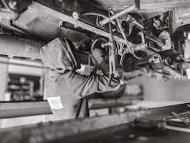A mechanic wearing a hooded sweatshirt works under a vehicle, performing maintenance or repairs. The setting is likely a workshop or garage. The scene is in black and white, emphasizing the components and mechanical parts of the vehicle's underside.