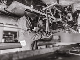 A mechanic wearing a hooded sweatshirt works under a vehicle, performing maintenance or repairs. The setting is likely a workshop or garage. The scene is in black and white, emphasizing the components and mechanical parts of the vehicle's underside.