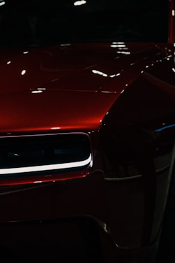 A close-up of a polished, shiny red car with a modern design. The focus is on the front section, highlighting a sleek headlight with LED lighting and reflecting surfaces creating an elegant look.