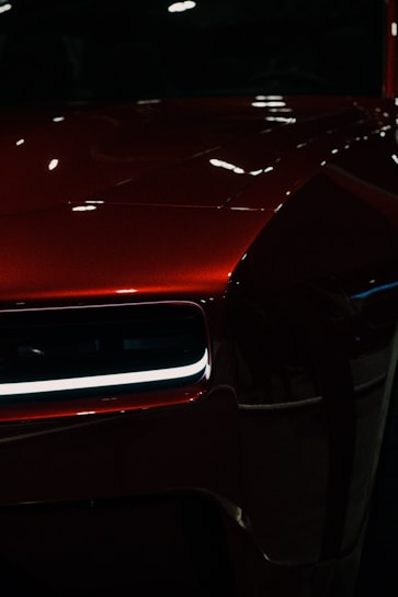 A close-up of a polished, shiny red car with a modern design. The focus is on the front section, highlighting a sleek headlight with LED lighting and reflecting surfaces creating an elegant look.