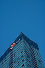 A tall, modern skyscraper with a glass facade dominates the scene, reflecting the clear blue sky. The building features the logo and name of a well-known hotel chain near the top, highlighting its corporate identity.