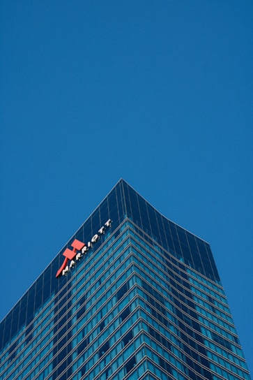 A tall, modern skyscraper with a glass facade dominates the scene, reflecting the clear blue sky. The building features the logo and name of a well-known hotel chain near the top, highlighting its corporate identity.