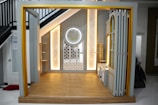 A small, elegantly designed prayer space under a staircase with warm lighting. The area features a wooden floor and a decorative wall panel with intricate patterns. There is a circular decoration on the wall with Arabic script.