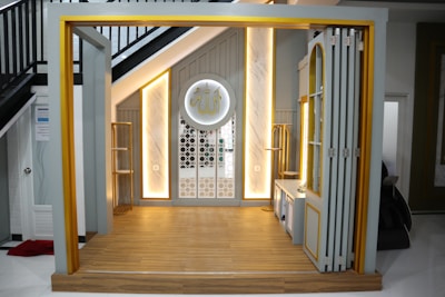 A small, elegantly designed prayer space under a staircase with warm lighting. The area features a wooden floor and a decorative wall panel with intricate patterns. There is a circular decoration on the wall with Arabic script.