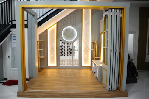 A small, elegantly designed prayer space under a staircase with warm lighting. The area features a wooden floor and a decorative wall panel with intricate patterns. There is a circular decoration on the wall with Arabic script.