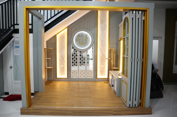 A small, elegantly designed prayer space under a staircase with warm lighting. The area features a wooden floor and a decorative wall panel with intricate patterns. There is a circular decoration on the wall with Arabic script.