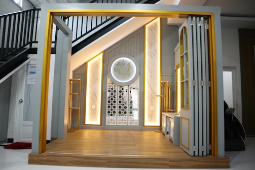 A modern interior with a small stage area, featuring warm lighting accents and a decorative Arabic calligraphy plaque on the wall. The space has a clean, minimalist design with gray and gold tones, complemented by wooden flooring and a staircase with black railings in the background.