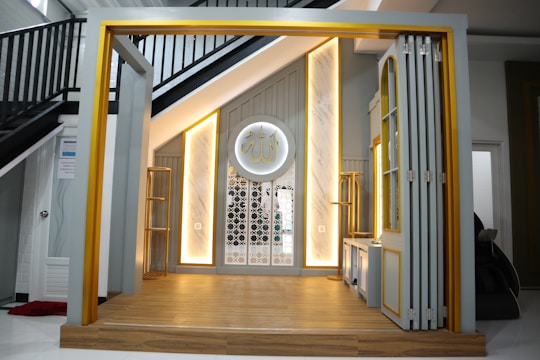 A modern interior with a small stage area, featuring warm lighting accents and a decorative Arabic calligraphy plaque on the wall. The space has a clean, minimalist design with gray and gold tones, complemented by wooden flooring and a staircase with black railings in the background.