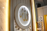 A beautifully decorated wall featuring a large circular plaque with ornate Arabic calligraphy in gold, surrounded by soft, ambient lighting. The background includes intricate white lattice patterns and vertical lines, creating an elegant and sophisticated atmosphere.