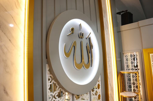 A beautifully decorated wall featuring a large circular plaque with ornate Arabic calligraphy in gold, surrounded by soft, ambient lighting. The background includes intricate white lattice patterns and vertical lines, creating an elegant and sophisticated atmosphere.