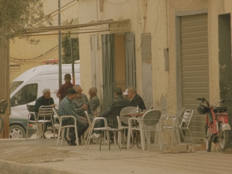 A group of elderly men are sitting around a table on a street corner, engaged in conversation. They are seated on metal chairs outside what appears to be a café. An individual stands nearby, while a red scooter and a white van are parked close by. The setting has a rustic, vintage atmosphere with muted colors and worn architecture.