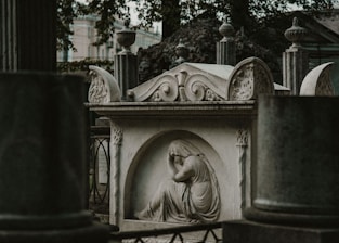 A stone sculpture depicting a somber, draped figure in an alcove, integrated into a decorative tomb structure. The surrounding environment includes metal railings and other stone elements with detailed carvings, suggesting a historical or memorial setting.