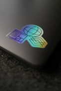 A holographic sticker placed on a sleek, dark surface. The sticker features geometric shapes including a globe, lines, and a diamond pattern, exhibiting a vibrant gradient effect.