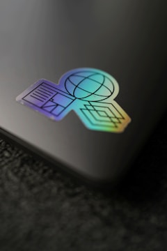 A holographic sticker placed on a sleek, dark surface. The sticker features geometric shapes including a globe, lines, and a diamond pattern, exhibiting a vibrant gradient effect.