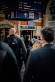 A crowded train station with people walking under a large sign displaying directions for buses, the underground, and taxis. The station has a classic architectural style with brick walls and arched doorways. The atmosphere appears busy with commuters wearing backpacks and coats.