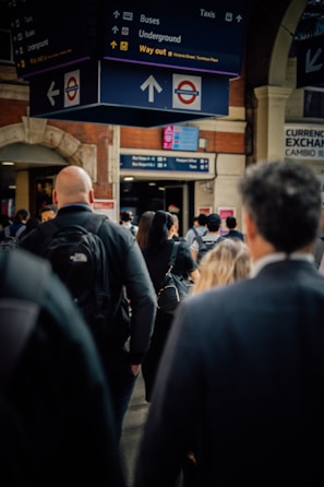 A crowded train station with people walking under a large sign displaying directions for buses, the underground, and taxis. The station has a classic architectural style with brick walls and arched doorways. The atmosphere appears busy with commuters wearing backpacks and coats.