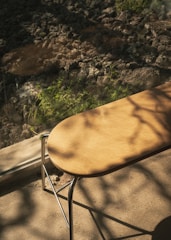 A wooden bench with metal legs is placed on a concrete surface, casting soft shadows. The background features a rocky terrain with patches of greenery and dappled sunlight creating an interplay of light and shadow.