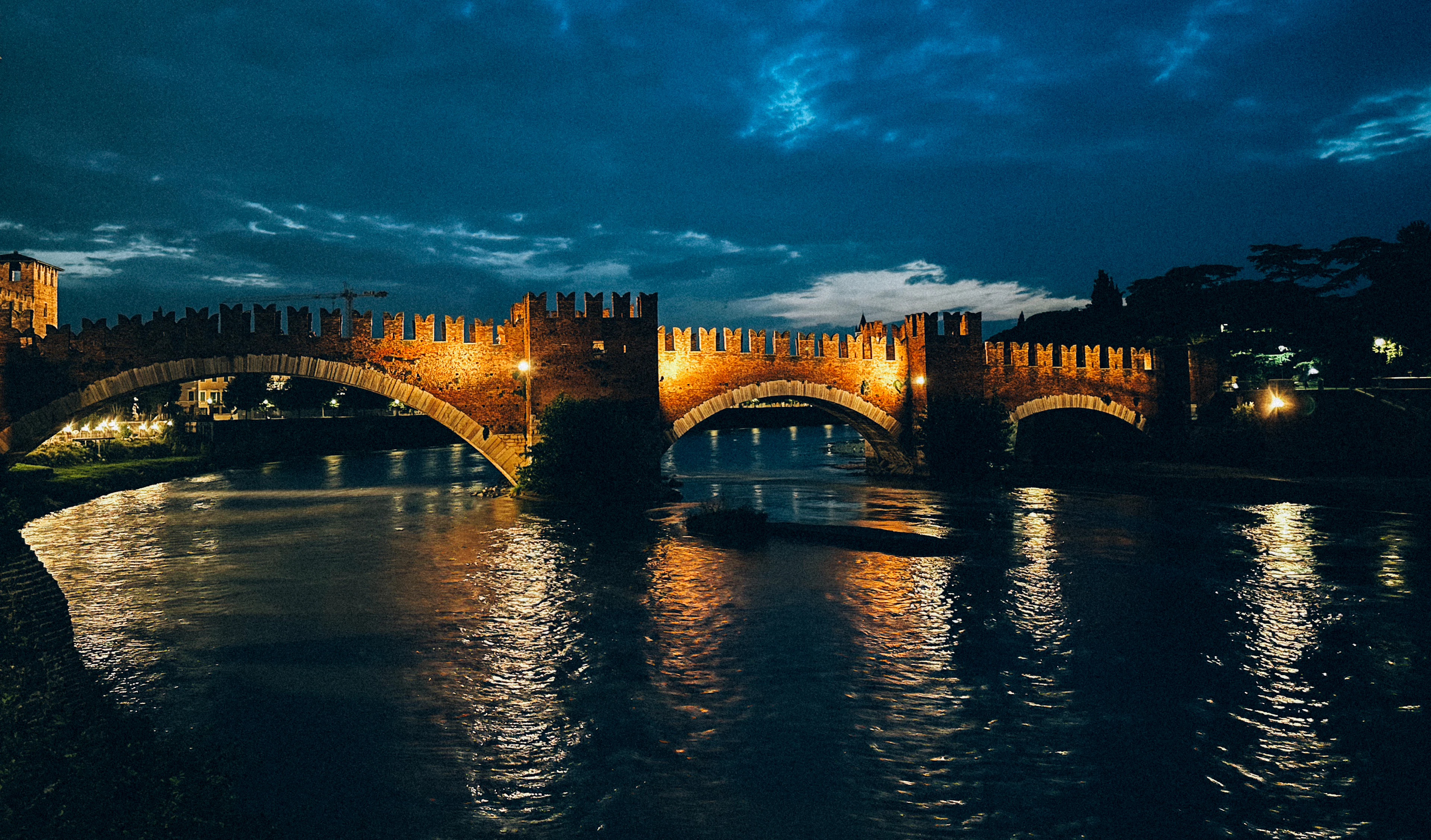 The swallowtail battlements and three brick arches of Verona's Ponte Scaligero floodlit gold at blue hour, the light running in long reflections down the dark Adige beneath streaked cloud.
