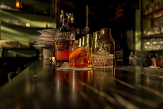 A dimly lit bar counter with a whiskey bottle, a cocktail glass garnished with an orange peel, and a glass decanter. The background is blurred, featuring shelves with various bottles. Warm lighting casts shadows, creating an intimate atmosphere.