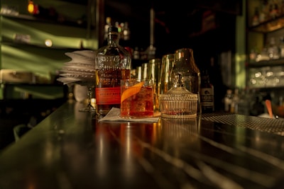 A dimly lit bar counter with a whiskey bottle, a cocktail glass garnished with an orange peel, and a glass decanter. The background is blurred, featuring shelves with various bottles. Warm lighting casts shadows, creating an intimate atmosphere.
