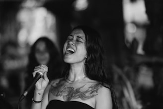 A woman passionately singing into a microphone, eyes closed, with a detailed bird tattoo on her chest. She has long, dark hair and is wearing a strapless top. The background is blurred, highlighting her expressive performance.