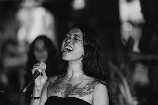A woman passionately singing into a microphone, eyes closed, with a detailed bird tattoo on her chest. She has long, dark hair and is wearing a strapless top. The background is blurred, highlighting her expressive performance.