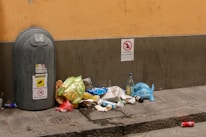 A street scene with a large gray trash bin and discarded garbage scattered on the pavement. The garbage includes plastic bags, bottles, paper, and a red aluminum can. Signage on the wall indicates restrictions regarding garbage disposal. The setting is urban, with a yellowish-beige wall and gray-tiled sidewalk.
