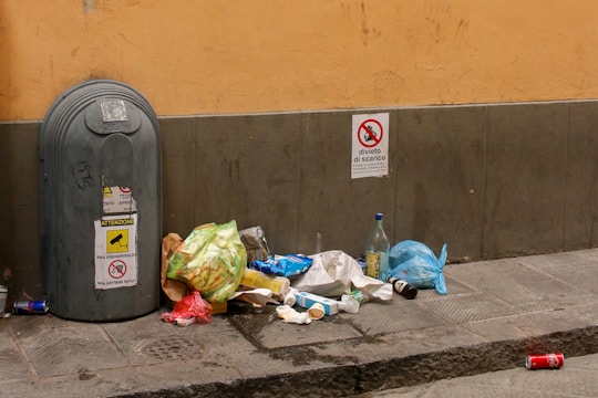 A street scene with a large gray trash bin and discarded garbage scattered on the pavement. The garbage includes plastic bags, bottles, paper, and a red aluminum can. Signage on the wall indicates restrictions regarding garbage disposal. The setting is urban, with a yellowish-beige wall and gray-tiled sidewalk.