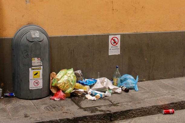 A street scene with a large gray trash bin and discarded garbage scattered on the pavement. The garbage includes plastic bags, bottles, paper, and a red aluminum can. Signage on the wall indicates restrictions regarding garbage disposal. The setting is urban, with a yellowish-beige wall and gray-tiled sidewalk.