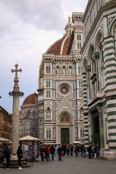 A large, ornate cathedral with a distinctive dome and intricate facade featuring Gothic architecture. The scene includes people gathered around a column with a cross on top, alongside street vendors and a display of postcards. The building's facade is adorned with geometric patterns in green, white, and pink tones.