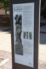 A vertical informational plaque is displayed outdoors, detailing the monument 'Kaddish' by Shelomo Selinger. The plaque contains a title, text in French, images of the sculpture, and Hebrew letters. The background shows a paved walkway, trees, and parts of a building.
